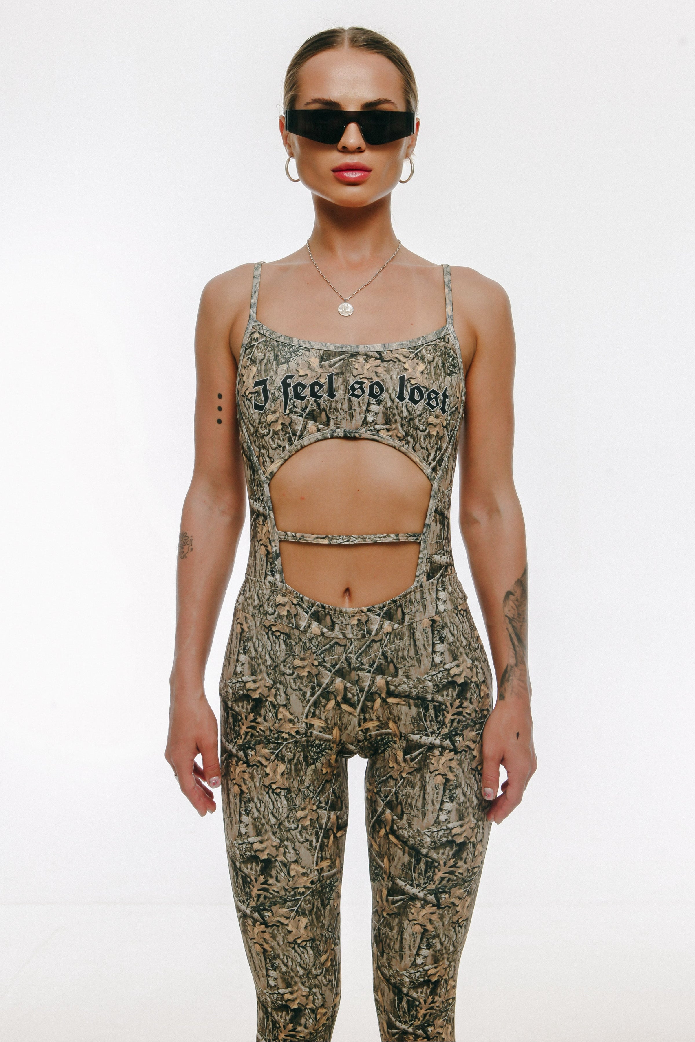 A model wearing a sleeveless camouflage jumpsuit with adjustable shoulder straps and a text graphic on the chest.